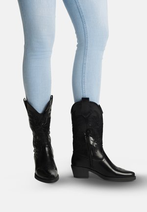 Person iført lyseblå skinny jeans og sorte broderede cowboystøvler med blokhæle, der går op midt på læggen, med den ene fod let løftet.