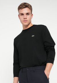 Crna dugorukava majica od pamuka, s malim zelenim Lacoste logom na prsima. Fit je opušten, s okruglim izrezom i cjevastim rukavima.