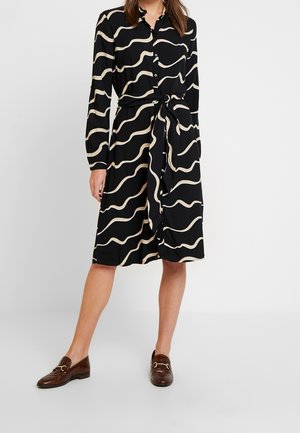 Frau trägt ein schwarzes Midikleid mit beigen wellenförmigen Streifen, einem gebundenen Taillengürtel, einer geknöpften Vorderseite und braunen Lederloafers.