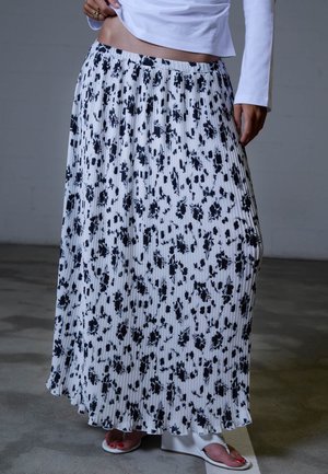 Mujer con falda maxi plisada blanca con estampado floral negro, top blanco de manga larga y sandalias de cuña blancas con esmalte de uñas rojo en los dedos.
