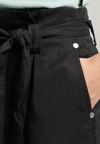 Pantaloni neri in tessuto con vita a cintura, tasche laterali e dettagli in metallo argentato. La texture è liscia con un design leggermente strutturato.