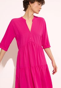 Fuksjowa sukienka z dekoltem w serek i krótkimi bufkami, z warstwowym designem i dwoma ozdobnymi guzikami przy dekolcie. Gładka faktura materiału.