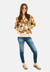 Gebreide trui in crème met bruine koeienprint, met lange mouwen en geribbelde manchetten. Gepaard met middelmatig gewassen jeans en sneakers.