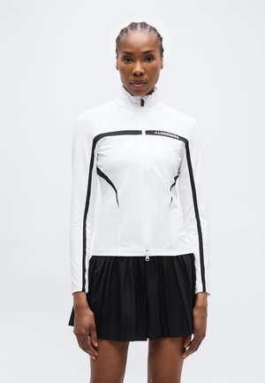 Γυναίκα με πλεκτά μαλλιά, που φοράει λευκό μπουφάν με μακριά μανίκια και φερμουάρ με μαύρες ρίγες και μαύρη πλισέ φούστα, όρθια μπροστά σε λευκό φόντο.