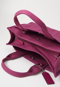 Fioletowa torba płócienna z dwoma uchwytami, zamykaną na zamek komorą i metką. Wnętrze jest przestronne z teksturowaną podszewką.