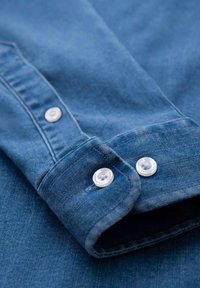 Denimhemdmanschette mit zwei klaren Kunststoffknöpfen, in mittlerem Blauton und subtiler Textur, die Nähdetails und Falten zeigt.