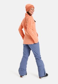 Fleece jack in lichtoranje met gestructureerde schouderpartijen, gecombineerd met blauwe sneeuwbroeken. Het model draagt een bijpassende oranje geribde beanie.