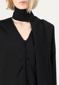 Blusa nera con righe verticali, caratterizzata da un colletto alto, maniche lunghe e chiusura a bottoni sul davanti. Tessuto liscio e leggero.
