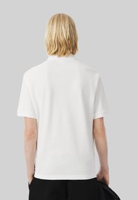 Person med axellångt blont hår iklädd en enkel vit kortärmad polo skjorta, stående mot en grå bakgrund.