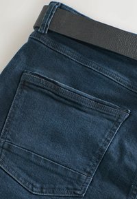 Tumed denimist valmistatud teksad tagataskuga ja must sile nahkrihm nähtav üleval. Kanga pind on sile ja kerge venivusega.