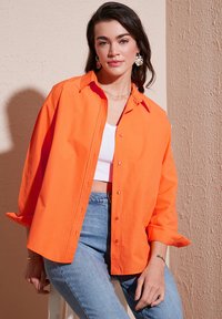 Felrood button-up shirt met een kraag, iets oversized pasvorm, lange mouwen opgerold, gecombineerd met een wit crop top en lichtblauwe jeans.