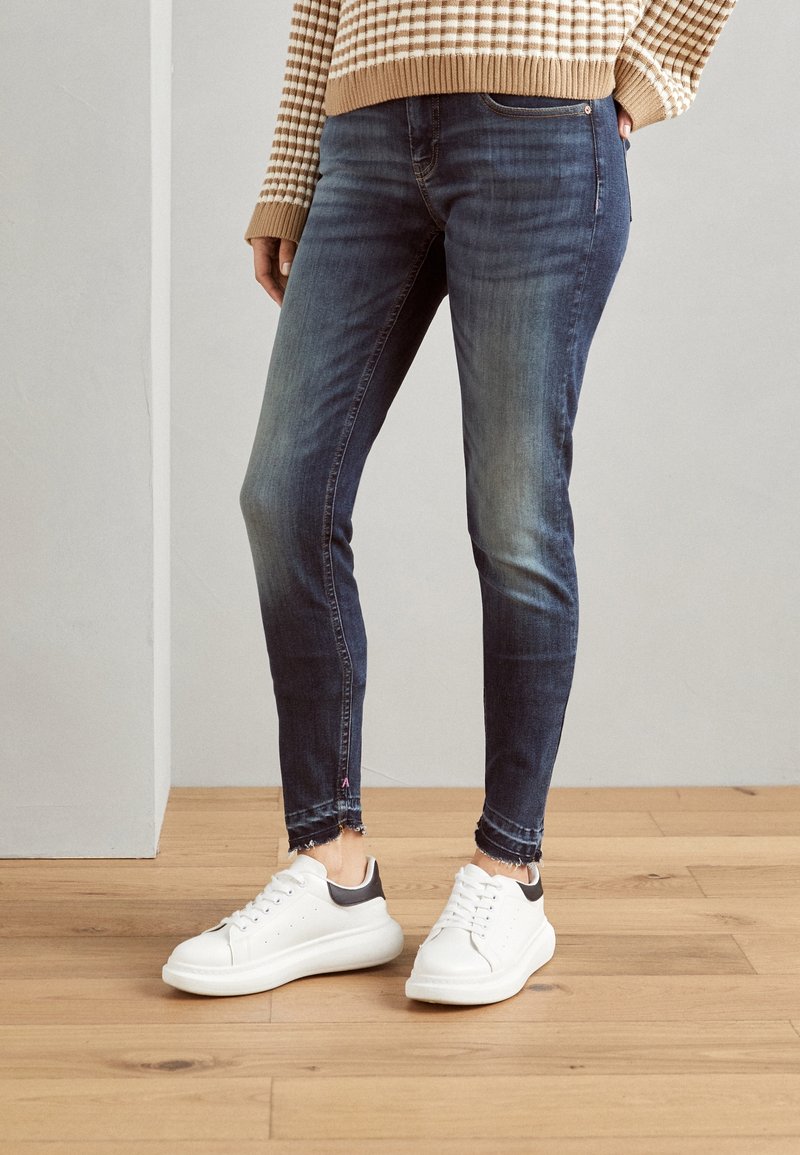 Vaqueros ajustados azules con dobladillos deshilachados, combinados con zapatillas blancas y negras. El suéter tiene rayas beige y blancas, y cuenta con un cuello acanalado.