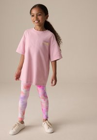 Ružové oversize tričko s krátkymi rukávmi a detailom loga. Farebné legíny s tie-dye vzorom v odtieňoch ružovej, fialovej a oranžovej. Viditeľné biele tenisky.