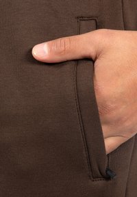 Brązowe dresowe spodnie z boczną kieszenią na czarny zamek błyskawiczny i prążkowaną fakturą. Ręka spoczywająca na kieszeni podkreśla design.