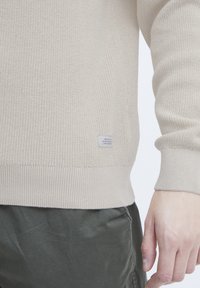 Pull en tricot beige avec un ourlet côtelé, présentant une petite étiquette en tissu sur le côté. La texture est lisse avec un motif de tissage lâche.