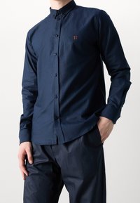 Mörkblå skjorta med knappar, tillverkad av slät tyg, med en smal krage, långa ärmar och en broderad orange "II"-detalj på bröstet.