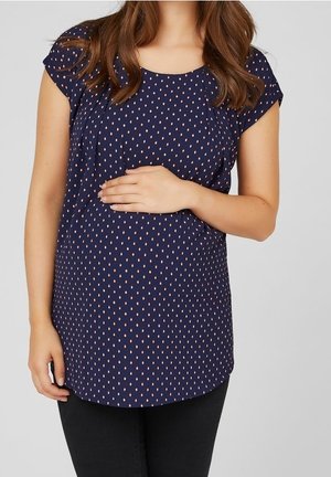 Kobieta ubrana w granatową bluzkę w groszki i czarne spodnie, trzymająca rękę na brzuchu w ciąży.