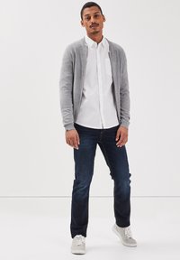 Szary kardigan na białej koszuli z guzikami, połączony z ciemnymi dżinsami i białymi trampkami. Gładka faktura, casualowy design i brak widocznych wzorów.