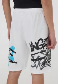 Pantaloni corti bianchi in cotone con una vestibilità rilassata. Presentano stampe in stile graffiti nere e blu sui lati. Fascia elastica in vita per il massimo conforto.