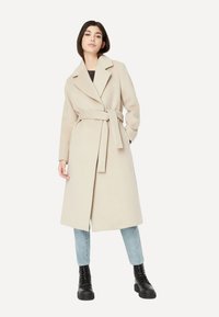 Beiger Wollmantel mit Gürtel, Reverskragen, langen Ärmeln und knielangem Design. Kombiniert mit hellblauen Jeans und schwarzen Kampfstiefeln.