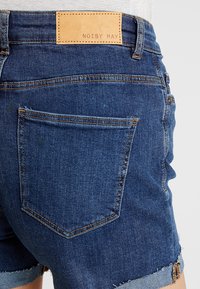 Džínové šortky tmavě modré barvy s hnědou koženou nášivkou s nápisem "NOISY MAY." Mají roztřepené lemy a klasický pětikapsový design.