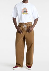 Hombre que lleva una camiseta blanca de Vans con un gráfico de computadora en llamas, pantalones marrones de pierna ancha con cordón blanco y zapatos de tela color burdeos.