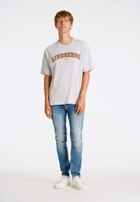 Grå bomullströja med "LINDBERGH 1927" tryckt i orange, tillsammans med blå jeans och vita sneakers. Modellen står framför en neutral bakgrund.