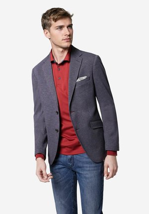 Blazer van textuur donkerblauwe stof met notch revers, zwarte knopen en een witte zakdoek, gecombineerd met een rode polo en denim.