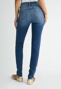 Jeans in denim blu slim-fit, con tasche posteriori cucite con un motivo geometrico e vita media, abbinati a scarpe piatte dorate.