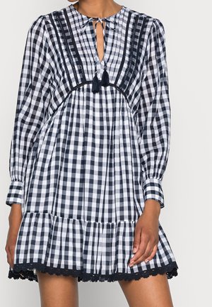 Mujer con vestido de manga larga de cuadros vichy en blanco y negro, con cuello con lazo de borlas y dobladillo con ribete de encaje.