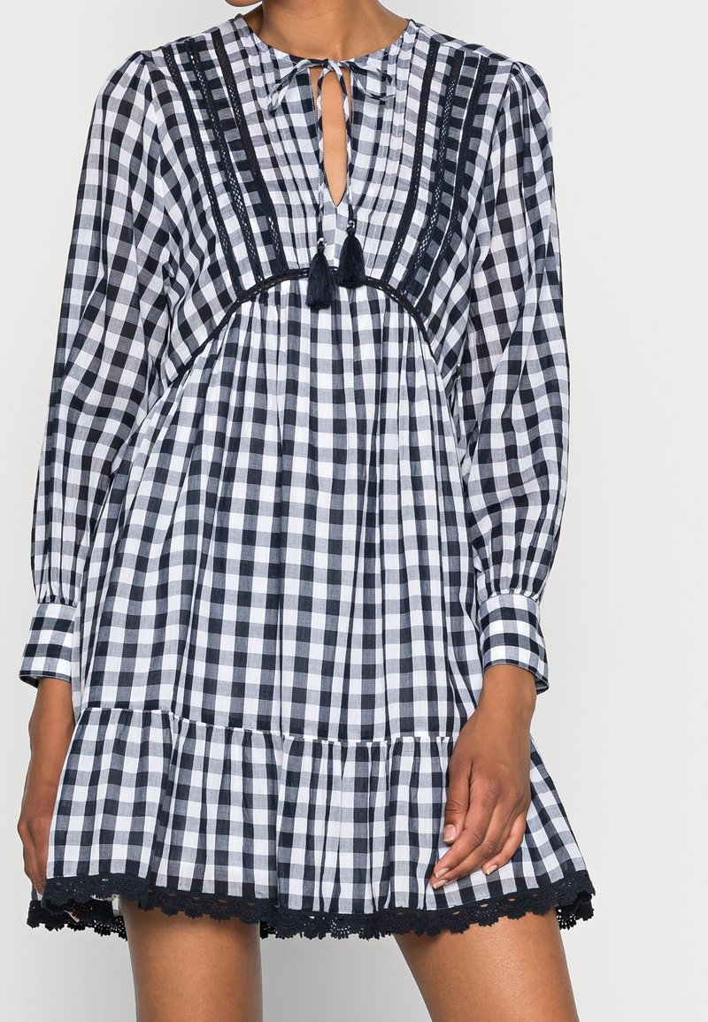 Femme portant une robe à manches longues à carreaux vichy noir et blanc, avec un col à lien à pampilles et un ourlet orné de dentelle.