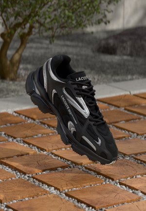 Baskets Lacoste noires et argent suspendues au-dessus de carreaux en bois en extérieur, avec du gravier et un petit arbre en arrière-plan.