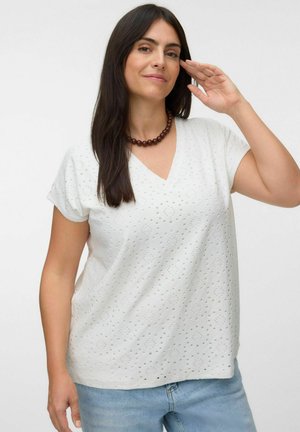 Femme aux longs cheveux foncés portant un T-shirt blanc à col en V ajouré, un jean bleu et un collier sombre en perles, levant la main droite près de son visage.