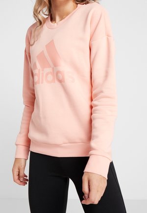 Kvinde iført en lyserød Adidas-sweatshirt med logo på brystet og sorte leggings, stående mod en ensartet lys baggrund.