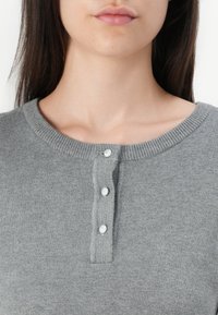 Pull en tricot gris avec un col rond côtelé et trois boutons blancs sur le devant, présentant une texture douce et un design ajusté.