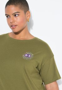 Osoba s krátkými vlasy, oblečená v zeleném tričku s fialovým a stříbrným designem oka, s zlatými kruhovými náušnicemi, se dívá mírně vlevo.