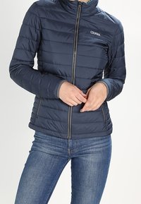 Marinblå puffjacka med hög krage, vertikalt kviltad design, dragkedja fram och en logotyp på övre vänstra sidan. Bärs med blå jeans.