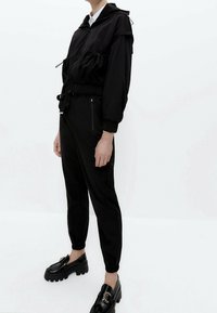 Persoon in een zwarte ritsjack met trekkoordzakken, bijpassende zwarte joggerbroeken en zwarte platform instappers met decoratieve gespen.