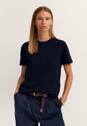 Woman with straight blonde hair wearing a black short-sleeve shirt, navy pants, and a brown braided belt with hands in pockets.