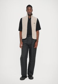 Ung man klädd i en beige zip-up väst över en svart kortärmad skjorta, svarta strukturerade byxor och svarta skor, stående mot en vit bakgrund.