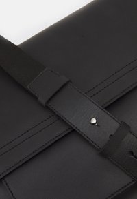 Borsa in pelle nera con una tracolla ampia e chiusura a bottone in argento su una superficie liscia, che mostra dettagliati punti di cucitura e texture.