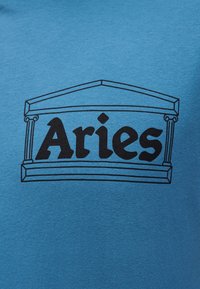 Μπλε ύφασμα με ασπρόμαυρο περίγραμμα γραφικού ελληνικού ναού που πλαισιώνει τη λέξη «Aries» με έντονη, κεφαλαία, με αγκύλες γραμματοσειρά.