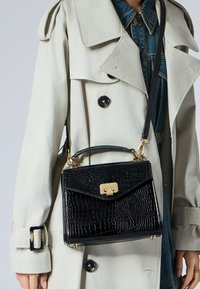 Borsa a tracolla in pelle nera con stampa coccodrillo, dotata di una chiusura dorata, manico corto e tracolla regolabile. Indossata con un trench coat chiaro.