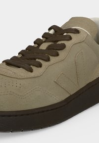 Baskets en daim beige avec bout perforé, lacets et semelle marron foncé, et logo cousu sur le côté.