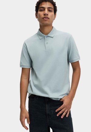 Junger Mann steht vor einem einfarbigen Hintergrund, trägt ein hellblaues Polohemd mit kurzen Ärmeln und schwarze Jeans, eine Hand in der Tasche.
