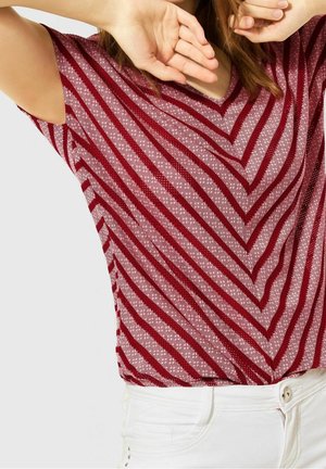 Person trägt ein rot-rosa gestreiftes V-Ausschnitt-Kurzarmoberteil, kombiniert mit weißer Hose, Arme erhoben und teilweise das Gesicht bedeckend.