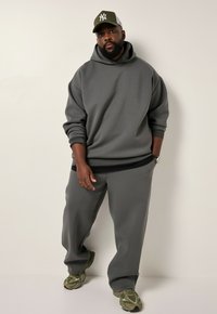 Grijze oversized hoodie met een trekkoord, gecombineerd met bijpassende losse sportbroeken. Draagt een olijfgroene pet en gemêleerde sneakers.