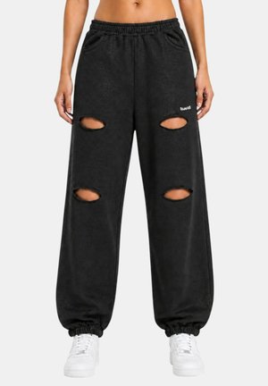 Pantalones deportivos negros con cintura y puños elásticos, con cuatro rasgaduras horizontales en las rodillas, usados con zapatillas blancas y un top corto.