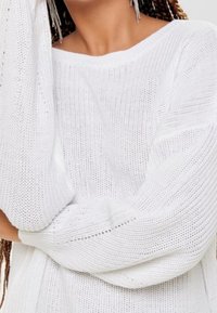 Pull en tricot blanc à coupe décontractée, avec des manches larges et un motif texturé. Le tissu semble doux et léger.