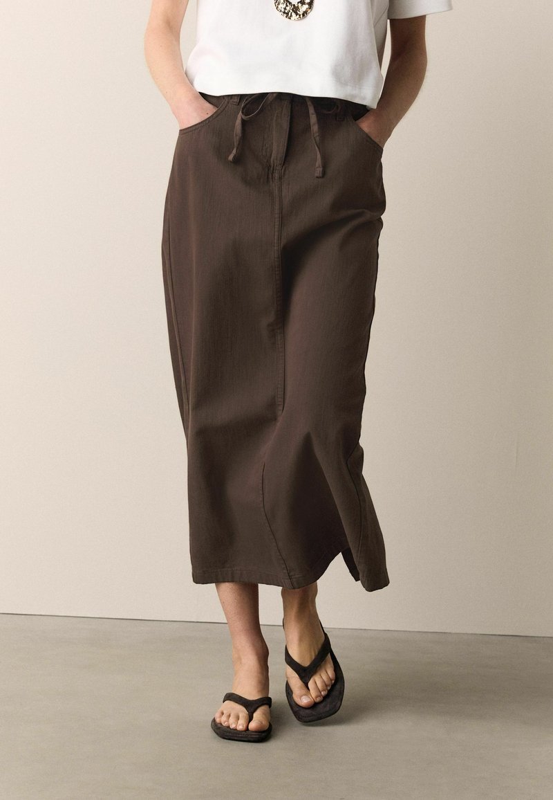 Woman wearing a brown midi skirt with front slit, white top, and black flip-flop sandals, standing with hands in pockets.